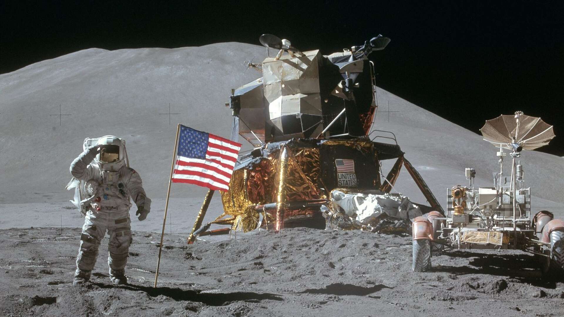 Jim Irwin gör honnör vid flaggan med månlandaren och månbilen och Mons Hadley i bakgrunden.