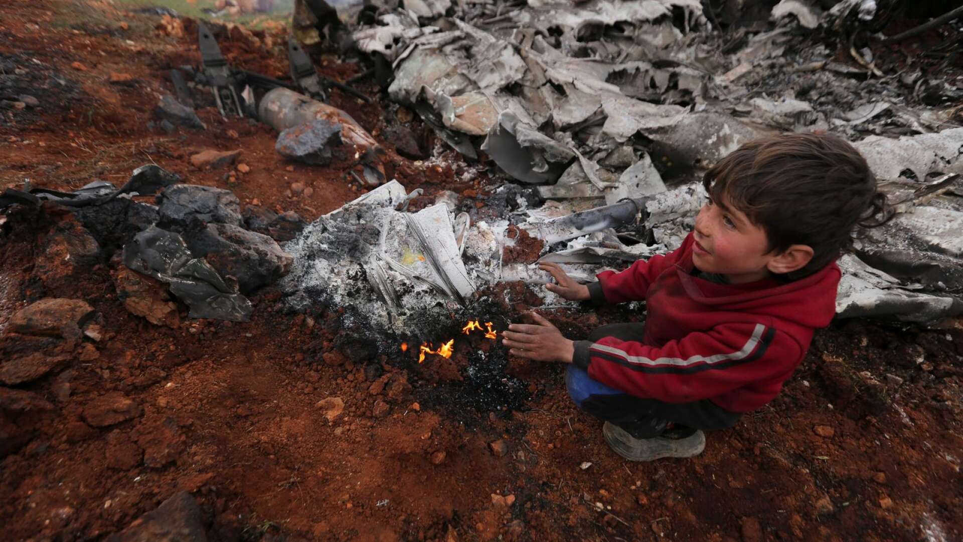 En syrisk pojke värmer sina händer vid ett brinnande vrak av en av regimens arméhelikoptrar utanför Aleppo i februari 2020.