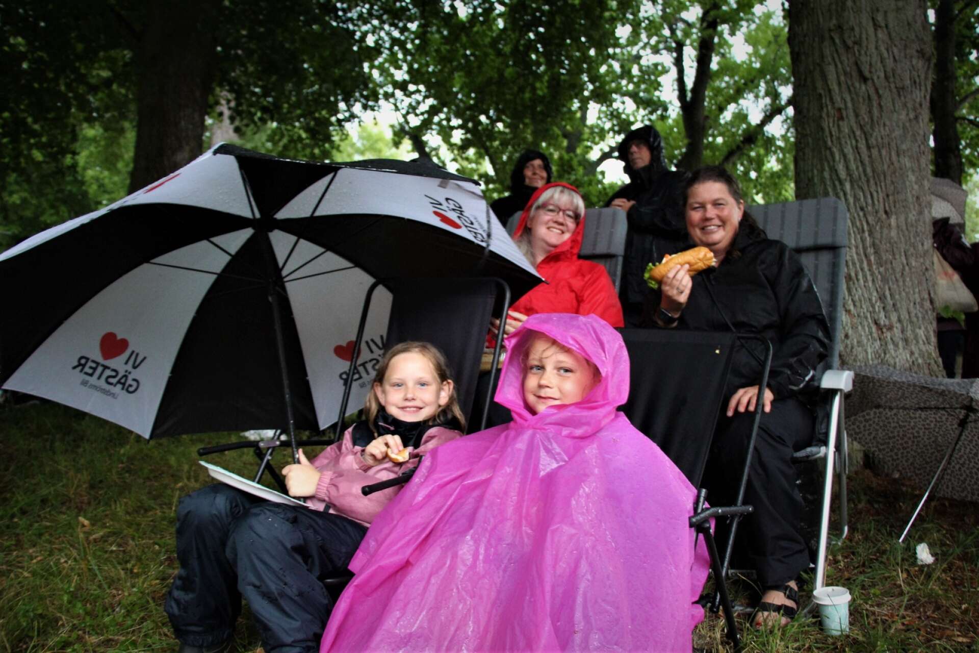 I rött sitter Jenny Seveborg med dottern Emelie framför. I svart sitter Maria Olofsson med dottern Julia Karlsson framför. Sällskapet äter glatt sitt fika. De har hittat regnskydd under ett av de stora träden.