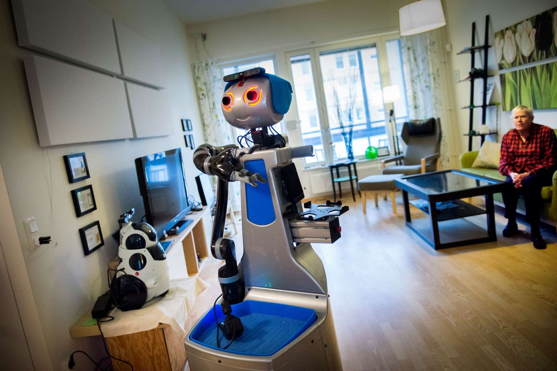 Bilden visar roboten Doro som hämtar medicin till Inge-Gerd Lindsten som väntar i soffan på Ängens forsknings- och innovationslägenhet i Örebro. Det här är ett exempel på hur robotar kan hjälpa till i vård och omsorgen. 