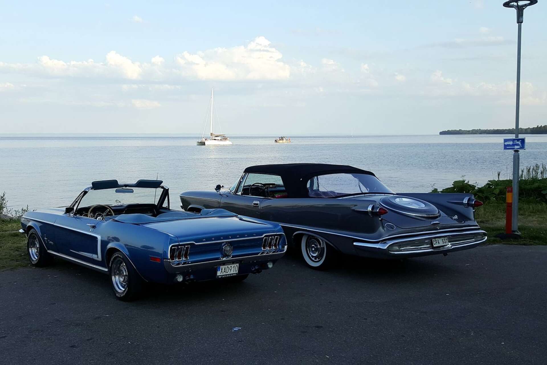 Ford Mustang av 1968 års modell och en Chrysler Imperial convertible av 1959 års modell parkerade i hamnen med vacker vy över Vättern.