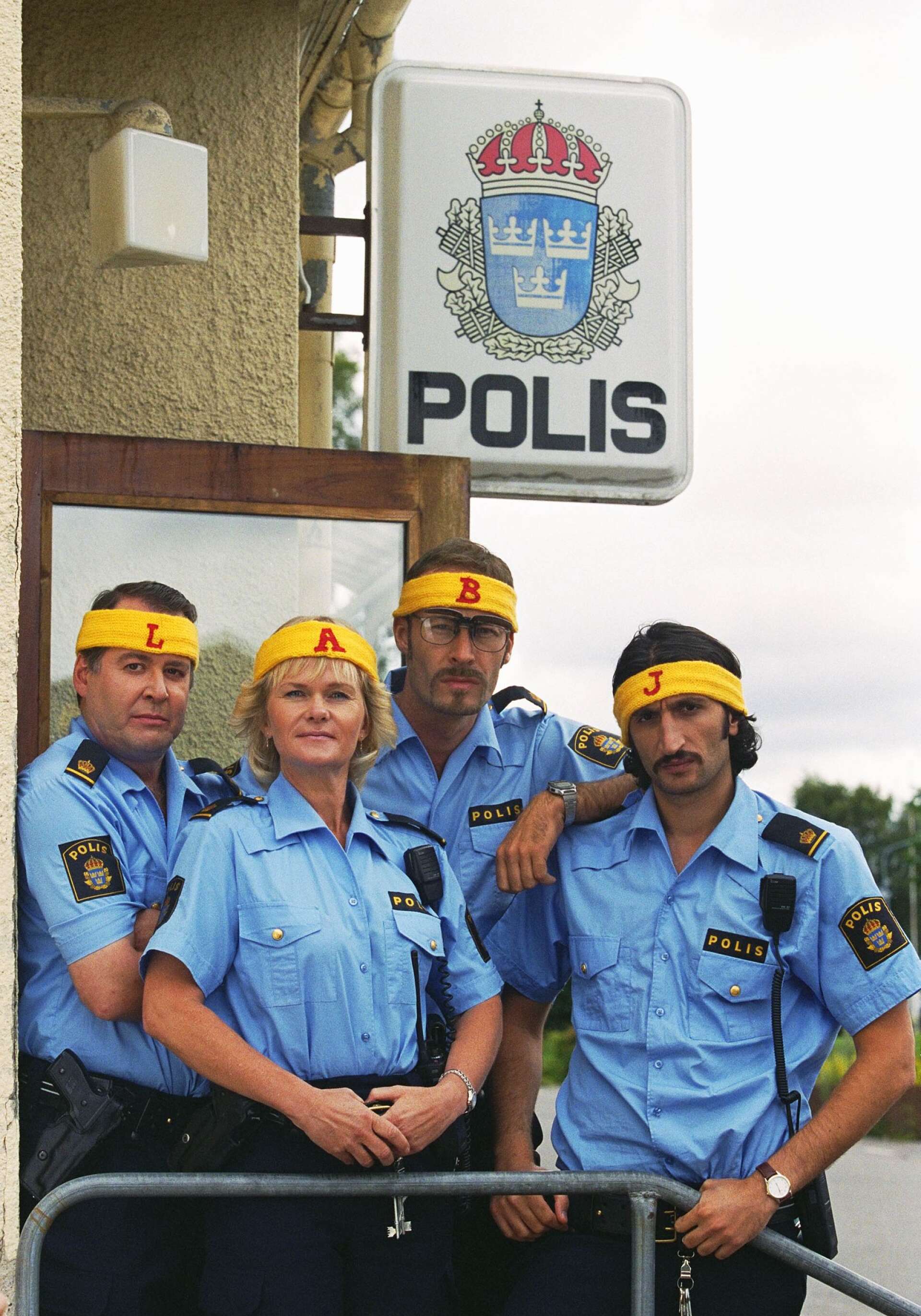 Skådespelarna utanför den nedläggningshotade polisstationen iförda de pannband som Benny (Torkel Petersson) stickat för landbandymatcherna. Från vänster: Göran Ragnerstam, Sissela Kyle, Torkel Petersson och Fares Fares.