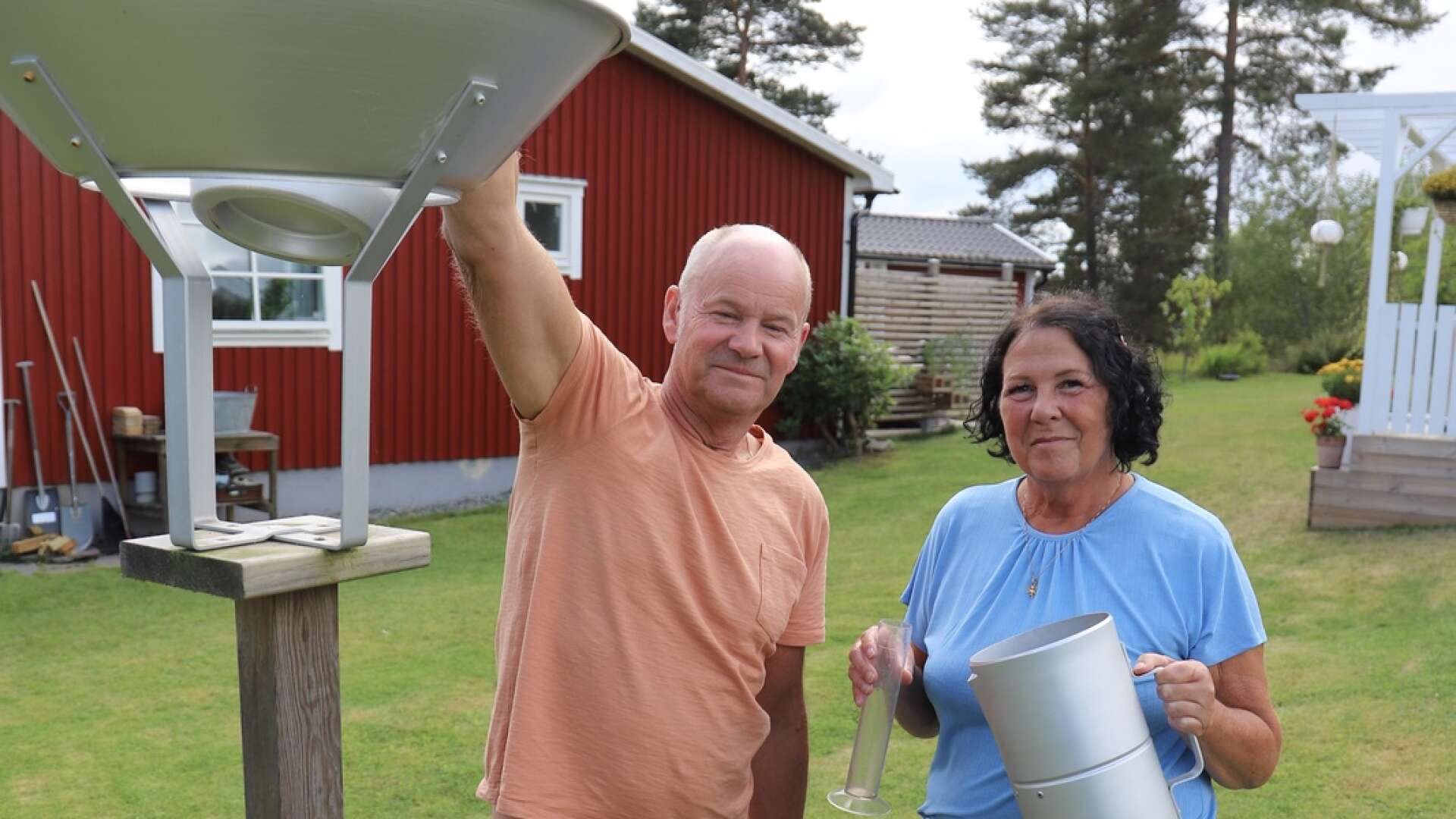 Vindskydd på en påle och en stor kanna får man av SMHI när man ska rapportera nederbörden. Här visar Kent och Catarina Erlandsson i Bengtsfors hur de arbetar.